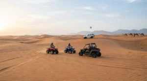 Helmeted riders on quad bikes and a buggy cross morning dunes, with a 4x4, a camel line and a balloon in the distance