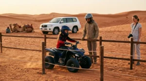 A helmeted child rides a small quad bike inside a fenced sand area as a guide and a parent watch, camels and a 4x4 behind